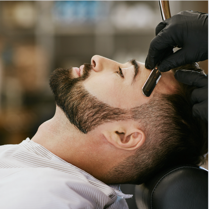 Professionel barbering med skin fade teknik, hvor en barber anvender en barberkniv til at forme håret på en kunde iført en stribet kappe i en moderne salon.